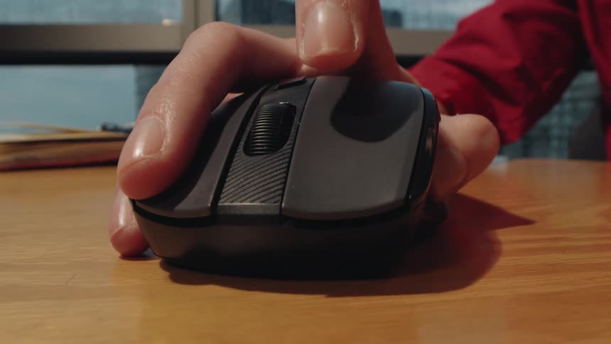 A close-up shot of a hand clicking a wireless computer mouse on a wooden desk by a window with a rainy city view