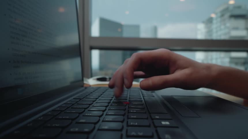 A close-up of a person's hand typing on a laptop keyboard in a modern office with a city skyline in the background