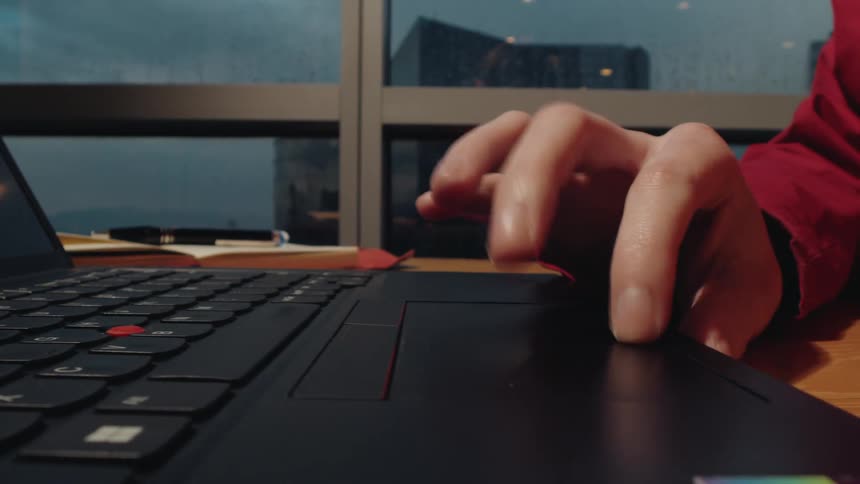 A close-up shot of a person's hands typing on a laptop keyboard and navigating with the trackpad in a dimly lit office