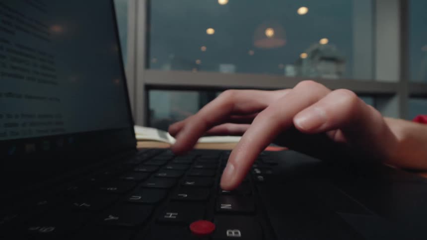 A close-up shot of a person's hands typing on a laptop keyboard in a modern workspace with a window view of the city