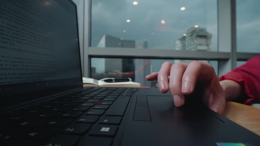 A focused close-up shows a hand navigating a laptop trackpad while working in a modern office overlooking the city