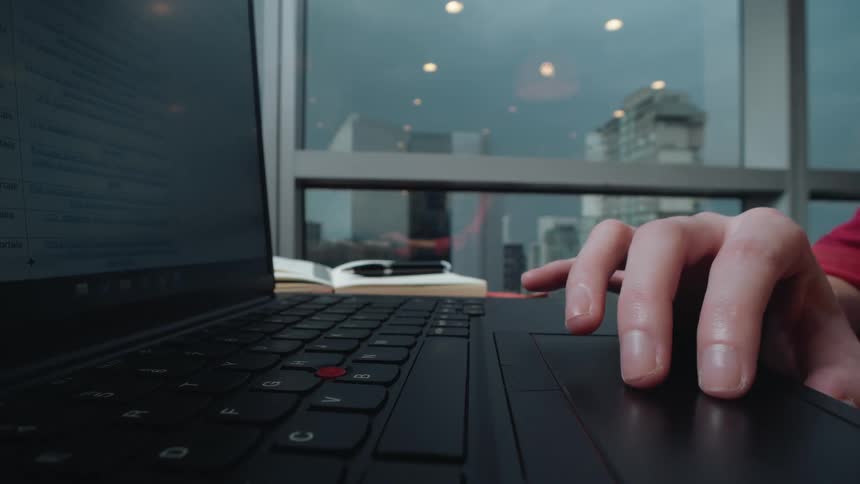 A person's hand scrolls on a laptop trackpad in a modern office with a city skyline visible through the window