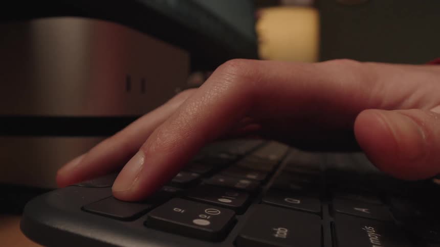 A close-up, low-angle view shows fingers typing frantically on a keyboard in a dimly lit room before pressing the escape key