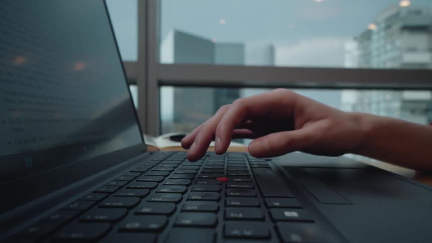A close-up of a person's hand typing on a laptop keyboard in a modern city office with a window view in the background