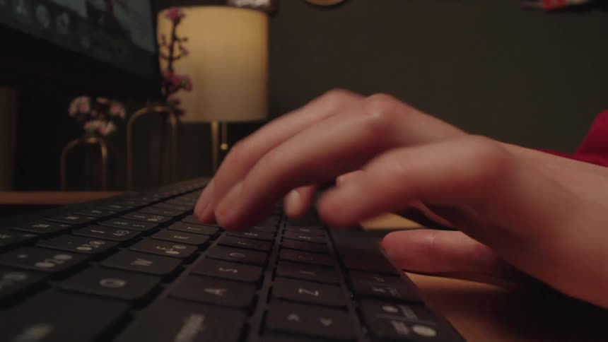 A side-view close-up shot of hands typing on a computer keyboard in a dimly lit room