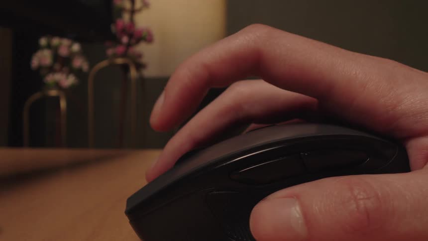 A close-up of a hand repeatedly clicking the button on a computer mouse while resting on a wooden desk