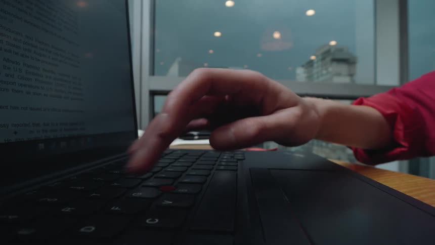 A low-angle close-up of a person's hand typing on a laptop keyboard with a city view from the window in the background