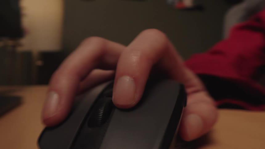 A person's hand rapidly clicking a computer mouse on a wooden desk in a close-up shot