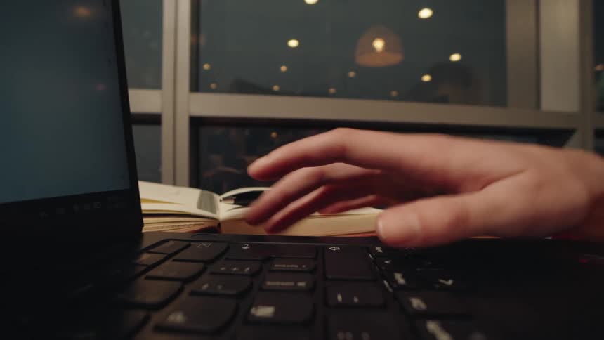 A person's hand types on a laptop keyboard in a dimly lit room, with a notebook and pen resting nearby