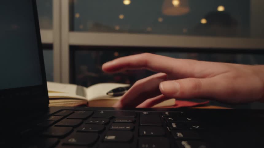 A close-up, slow-dolly shot of a hand typing on a laptop keyboard at a desk in the evening