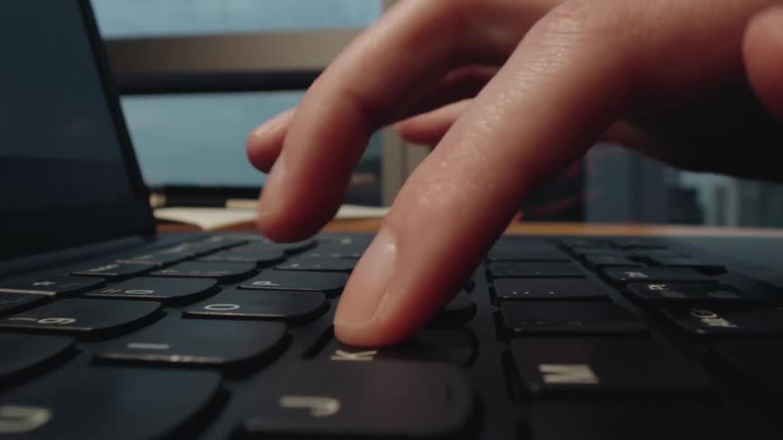 A low-angle macro shot dollies along a laptop keyboard as a person's fingers type in a modern office