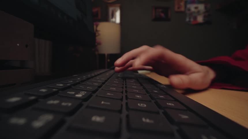 A dramatic close-up shot of a person's hands typing on a computer keyboard in a dimly lit room at night
