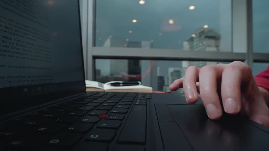 A close-up shot of a person's hand using the trackpad on a laptop to scroll through a document in an office with a city view