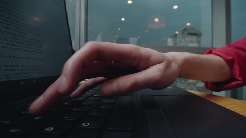 A close-up shot of a person's hands using a laptop's trackpad and keyboard to work in an office with a city view
