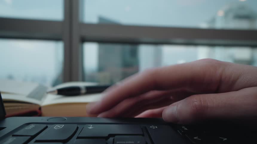 A close-up of a person's hand using the trackpad on a laptop in an office with a city view in the background