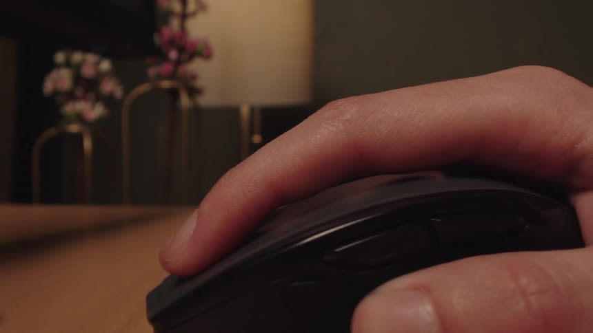 Extreme close-up of a person's hand operating a black wireless computer mouse on a wooden desk