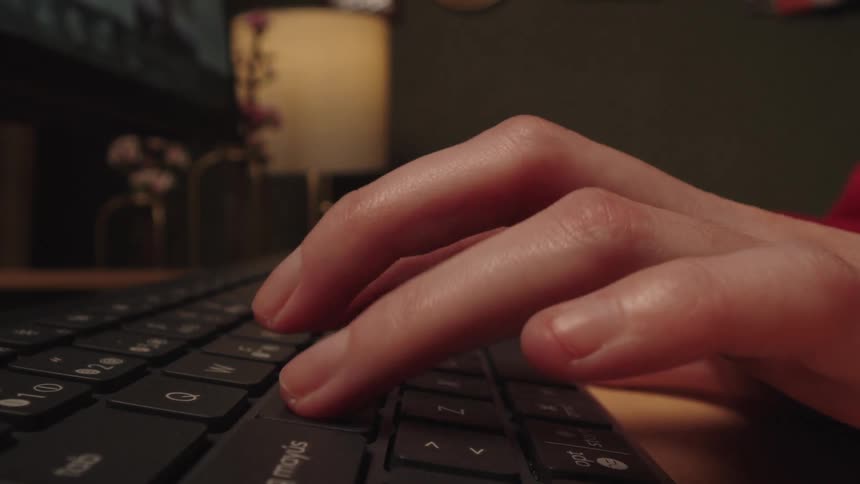 Extreme close-up of fingers typing on a black keyboard on a desk in a dimly lit room