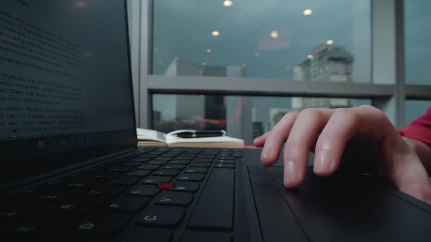 Close-up of a person's hand typing and using the trackpad on a laptop in a modern office with a city view in the background