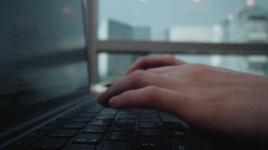 A close-up side view of a person's hands typing on a laptop keyboard in a modern office