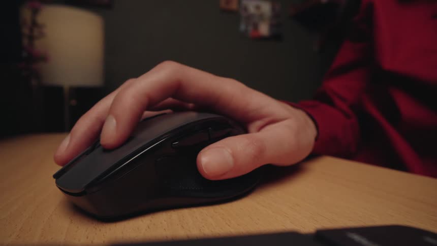 Static close-up shot of a man's hand clicking a wireless computer mouse on a wooden desk in a dark room