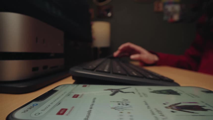 A low-angle shot shows a user scrolling through an online shopping website on a smartphone, with a keyboard and computer in the background