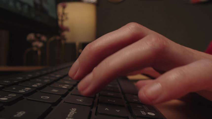 An extreme close-up side view of a woman's hands typing on a computer keyboard in a dimly lit room