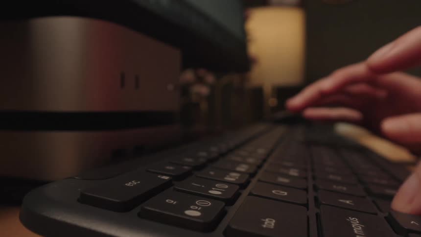 A close-up, low-angle shot of a person's hands typing on a computer keyboard in a dimly lit room