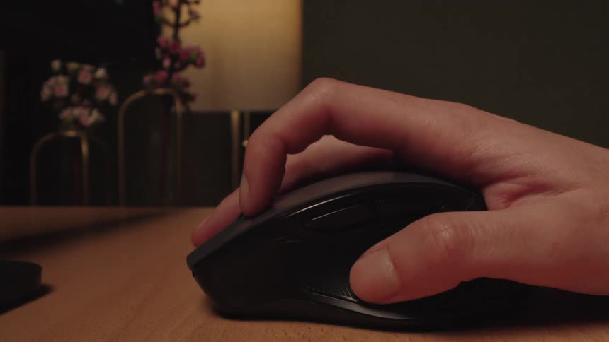 Close up of a hand on a wireless computer mouse, clicking and scrolling on a wooden desk in a dimly lit office