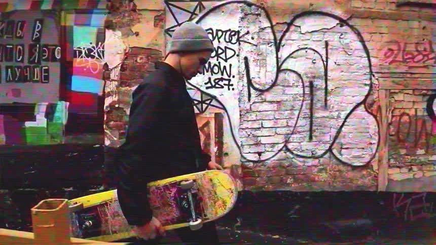 A young man with a skateboard walks in slow motion past a graffiti-covered brick wall with a retro VHS glitch effect