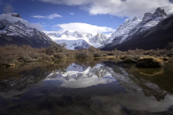 A breathtaking view of a snow-covered mountain range and cloudy blue sky perfectly mirrored in the still waters of a tranquil lake