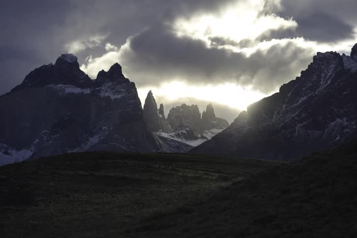 A time-lapse shows dramatic dark clouds rolling over a jagged, snow-capped mountain range as the sun shines through