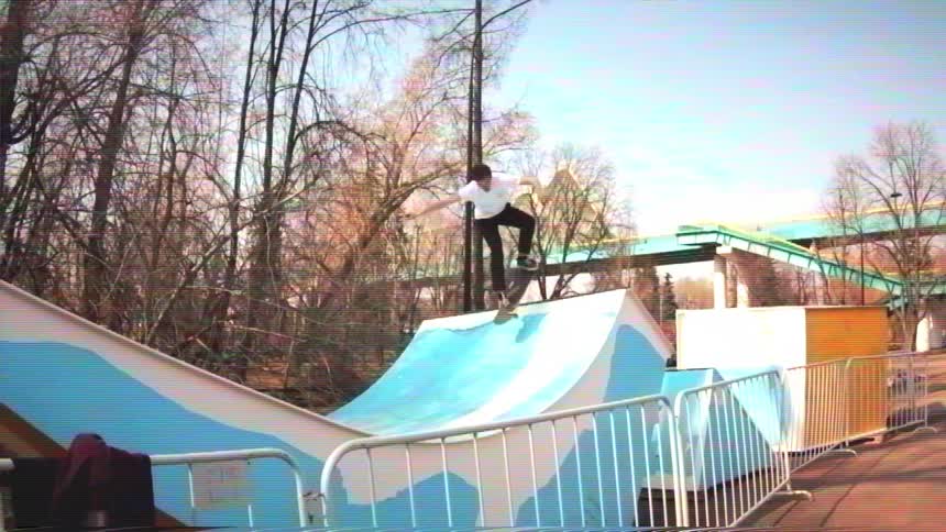 A young man performs a trick on a skateboard ramp in a park, captured in a slow-motion, vintage-style shot