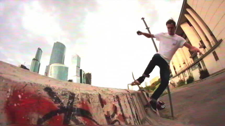A young man performs a slow-motion skateboard grind on a graffiti-covered concrete barrier with a modern city skyline in the background