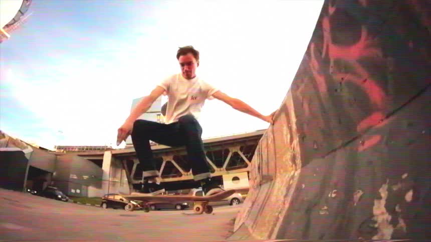 Slow motion VHS-style footage of a skateboarder performing an aerial trick on a concrete ramp under a city bridge