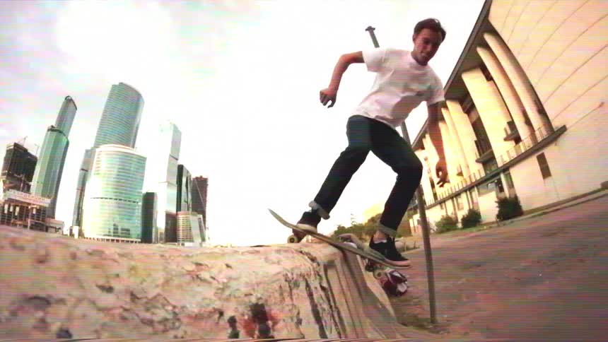A young man performs a skateboard grind on a graffiti-covered concrete ledge in slow motion, with a modern city skyline in the background