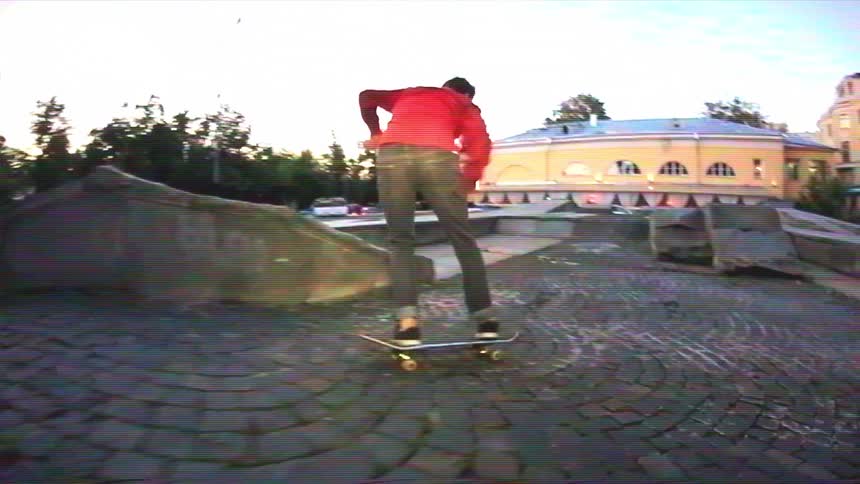A slow-motion, wide-angle shot captures a skateboarder in a red shirt doing a flip trick off a concrete ledge onto a cobblestone street