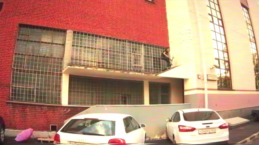 A man on a scooter performs daring balancing tricks on a narrow ledge of a red brick building