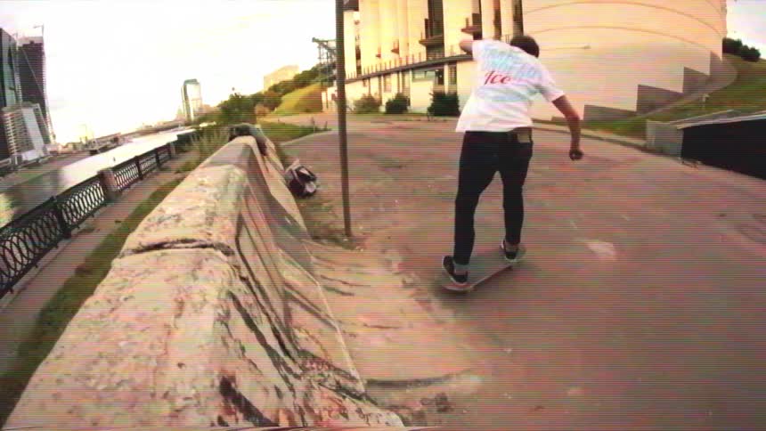 A young man performs a skateboarding trick on a concrete ramp along a riverfront with a modern city skyline in the background