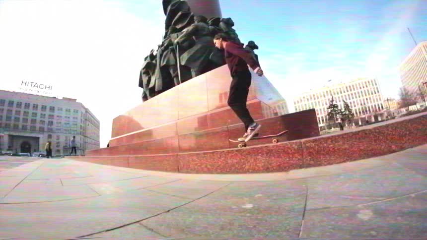 A skateboarder performs a complex flip trick onto the curved ledge of a city monument in a slow-motion, fisheye shot