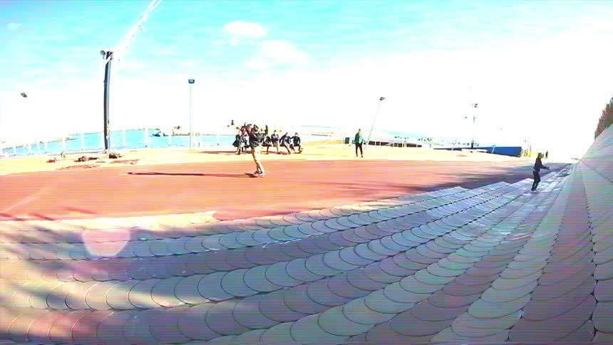 A skateboarder performs a jump trick in slow motion on a unique scalloped ramp near the waterfront on a sunny day