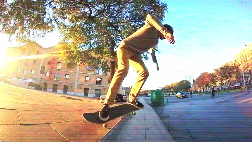 A skateboarder performs a tailslide to shove-it trick off a stone ledge in a sunny city plaza in slow motion
