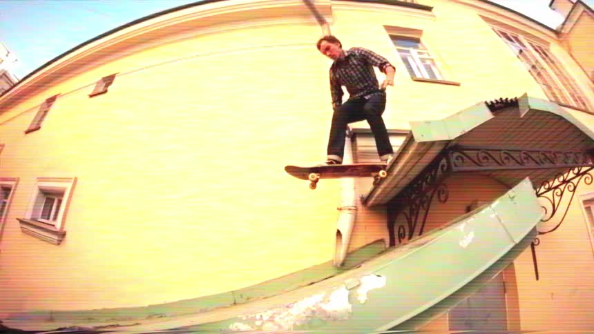 A skateboarder in slow motion climbs onto a roof, then drops in to ride down a curved metal awning in a gritty urban alley