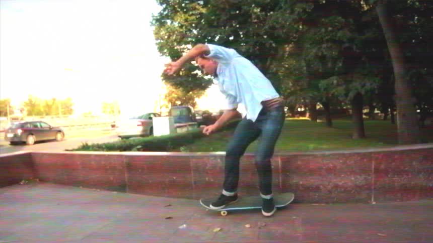 A skateboarder in a white shirt and dark jeans grinds along a stone ledge in slow motion with a VHS filter