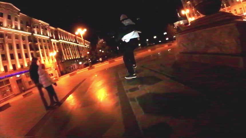 A skateboarder cruises through a brightly lit city plaza at night in a slow-motion, fisheye perspective