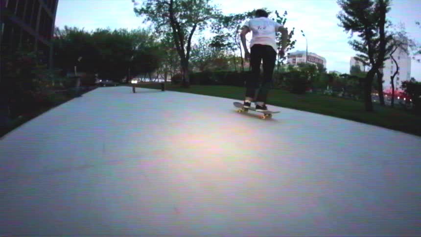 Slow motion shot of a skateboarder doing a pop shuvit and ollieing over a bench in a city park at dusk