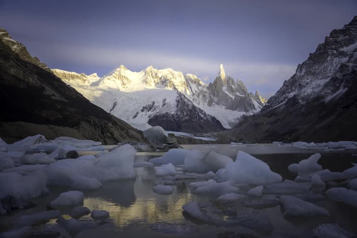 A majestic time-lapse captures the sunrise illuminating the snowy peaks of Cerro Torre, with its reflection in a glacial lake filled with icebergs