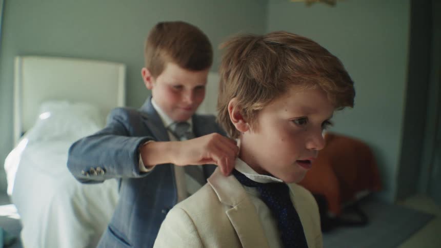 An older brother carefully helps his younger brother get dressed in a suit inside a bedroom