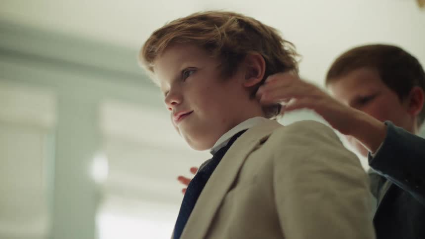 A young boy helps his brother or friend adjust his suit collar as they get dressed for a special event