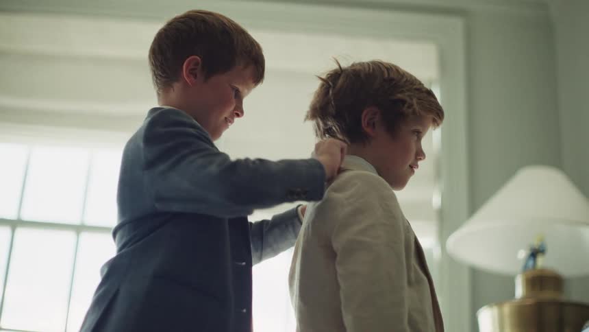 In a sweet and caring moment, a young boy adjusts his brother's collar as they get dressed in suits for a special event