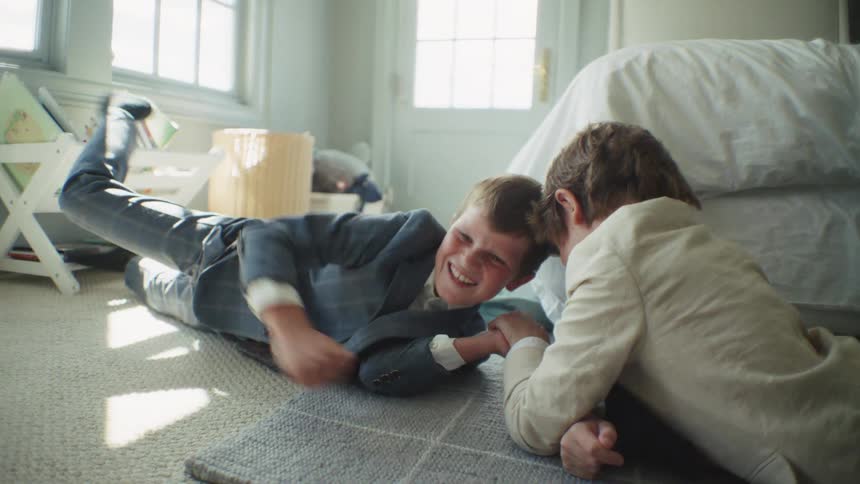 Two laughing brothers in suits playfully arm wrestle on the floor of their sunlit bedroom